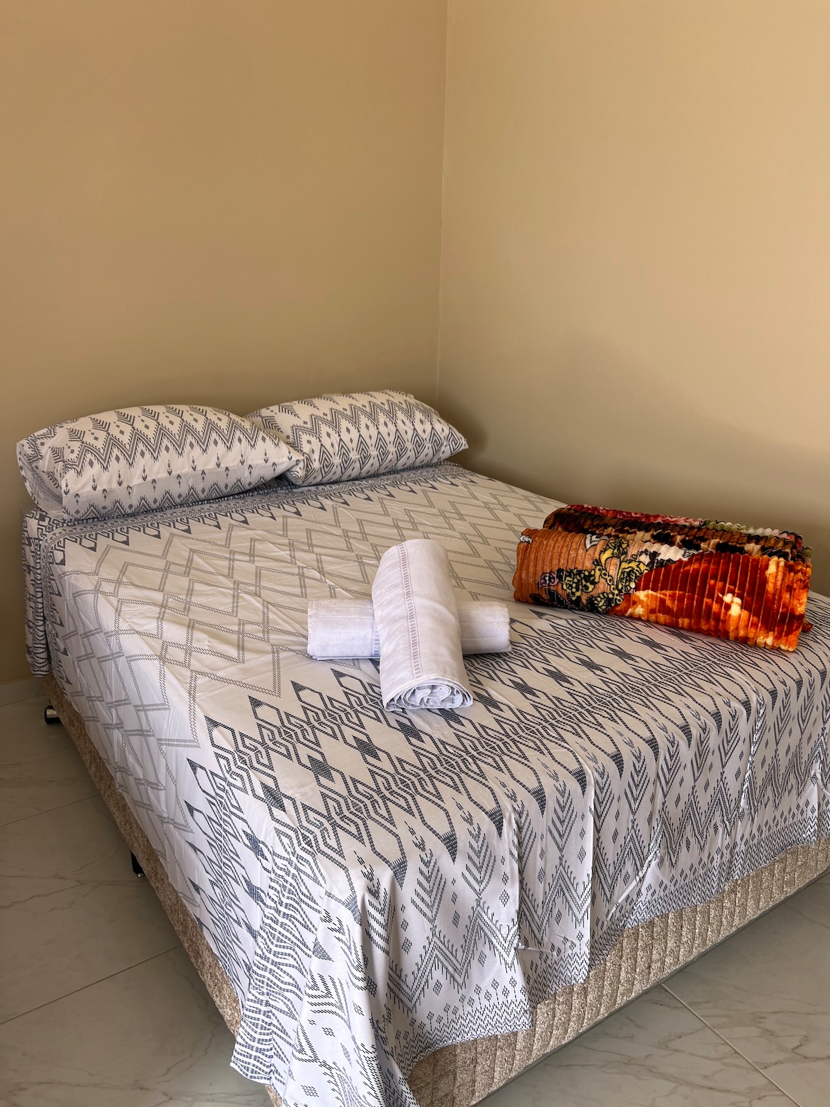 A spacious bed is dressed in a patterned white and gray duvet, complemented by two matching pillows. A rolled towel is placed neatly on the bed, along with an orange and colorful blanket, adding a layer of comfort to the inviting sleeping space.