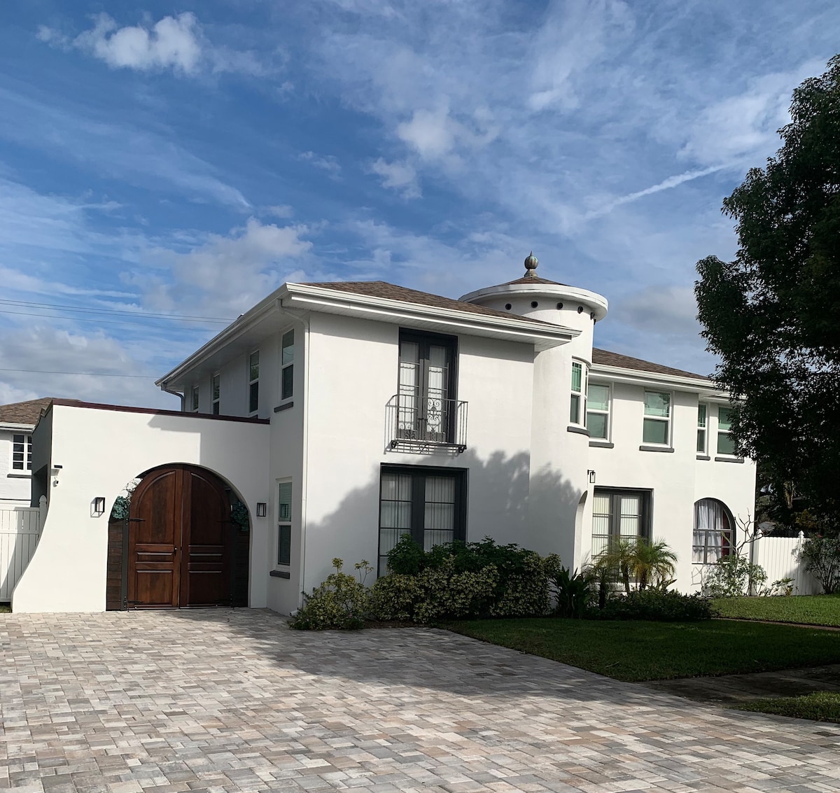 A modern two-story house is displayed under a partly cloudy sky. The white exterior features large windows and a distinctive circular turret. A textured stone driveway leads to a grand wooden door, providing an entry point to the welcoming residence.