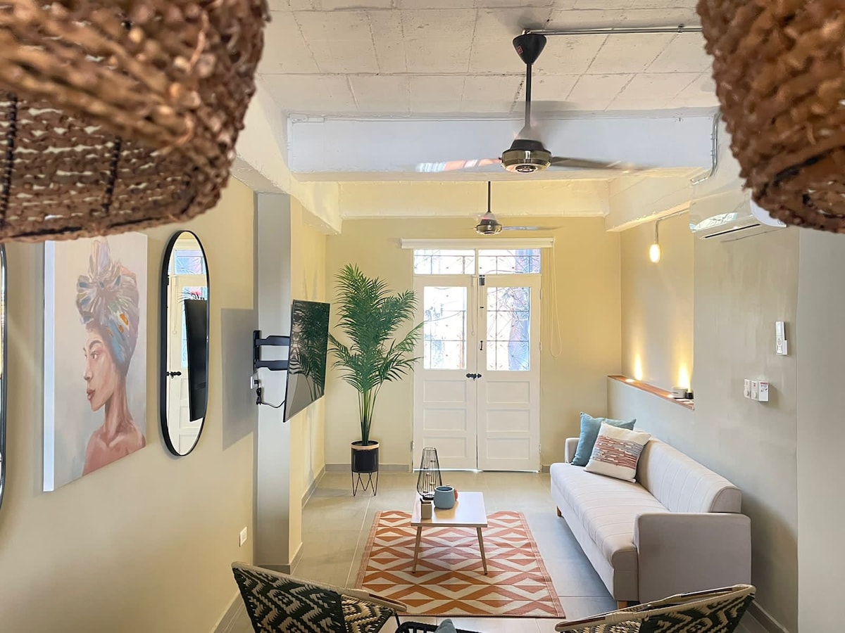 The living area features a light-colored sofa complemented by textured throw pillows. A medium-sized coffee table sits on a patterned rug, while a decorative plant adds a touch of greenery. Natural light fills the space through a large window, and stylish light fixtures enhance the ambiance.