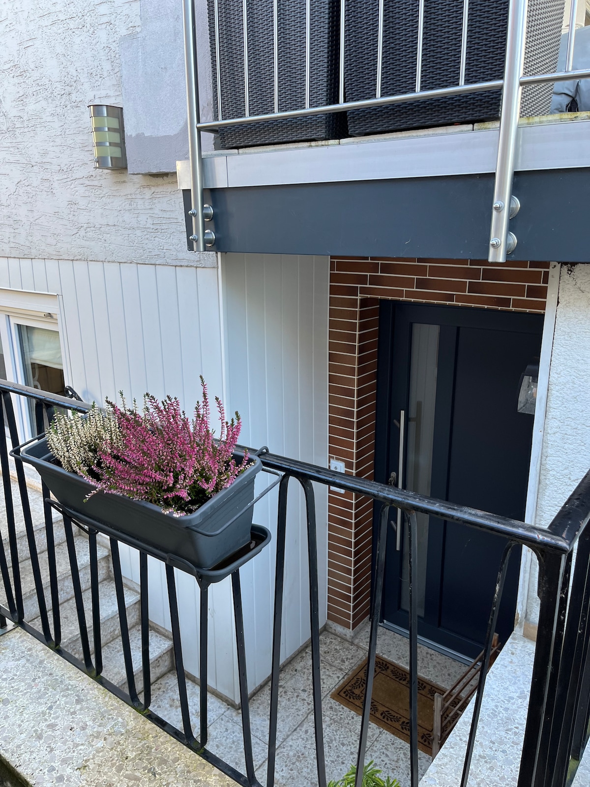 A ground-level entrance is framed by a dark door and features a planter box filled with vibrant heather plants. A metal railing adds a touch of elegance, while the textured wall and warm-toned bricks create a welcoming exterior.