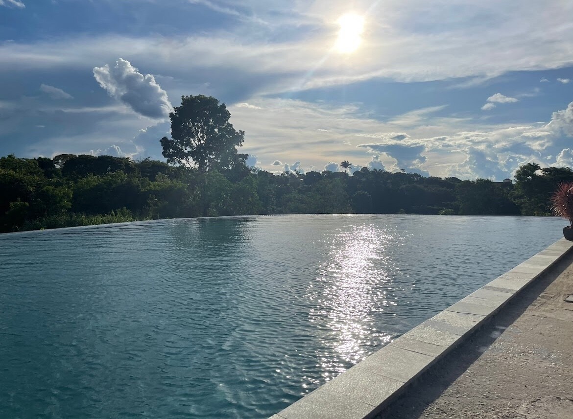 An infinity pool reflects the sunlight, creating a shimmering effect on the water's surface. Lush greenery is visible in the background, providing a serene natural setting. The sky features scattered clouds, enhancing the peaceful atmosphere of the outdoor space.