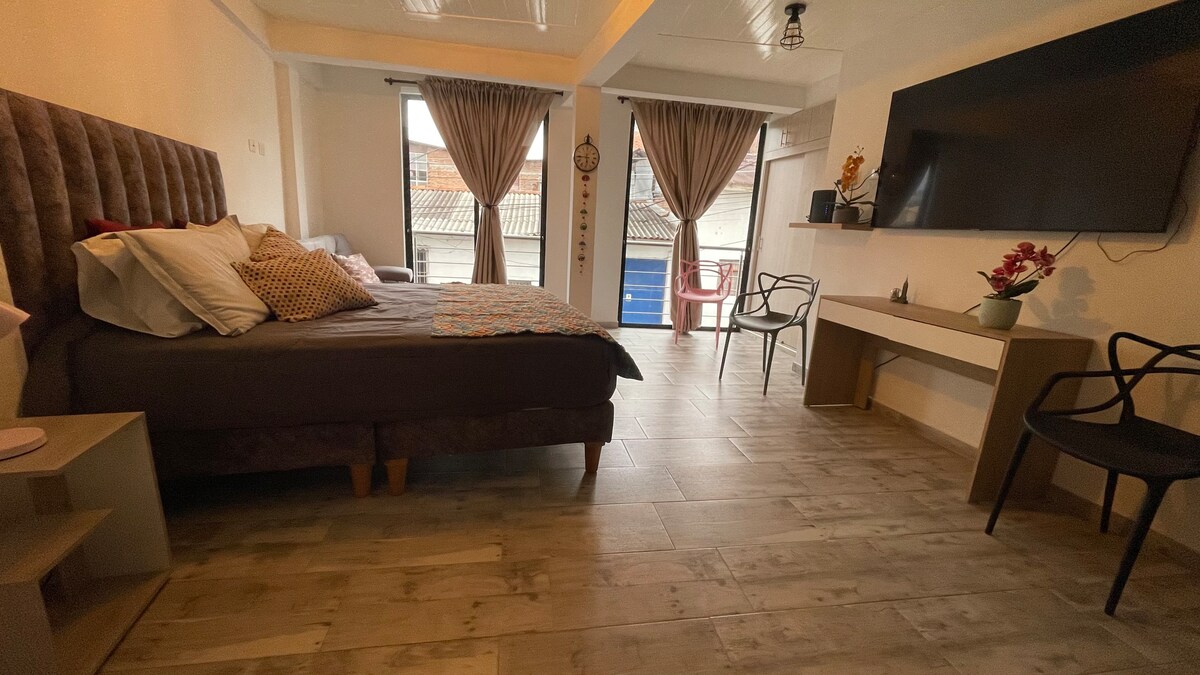 A spacious room is presented with a plush headboard supporting a large bed dressed in neutral linens. Two windows are adorned with soft curtains, allowing natural light to fill the room. A desk and chair are positioned near the wall, complemented by a seating area.