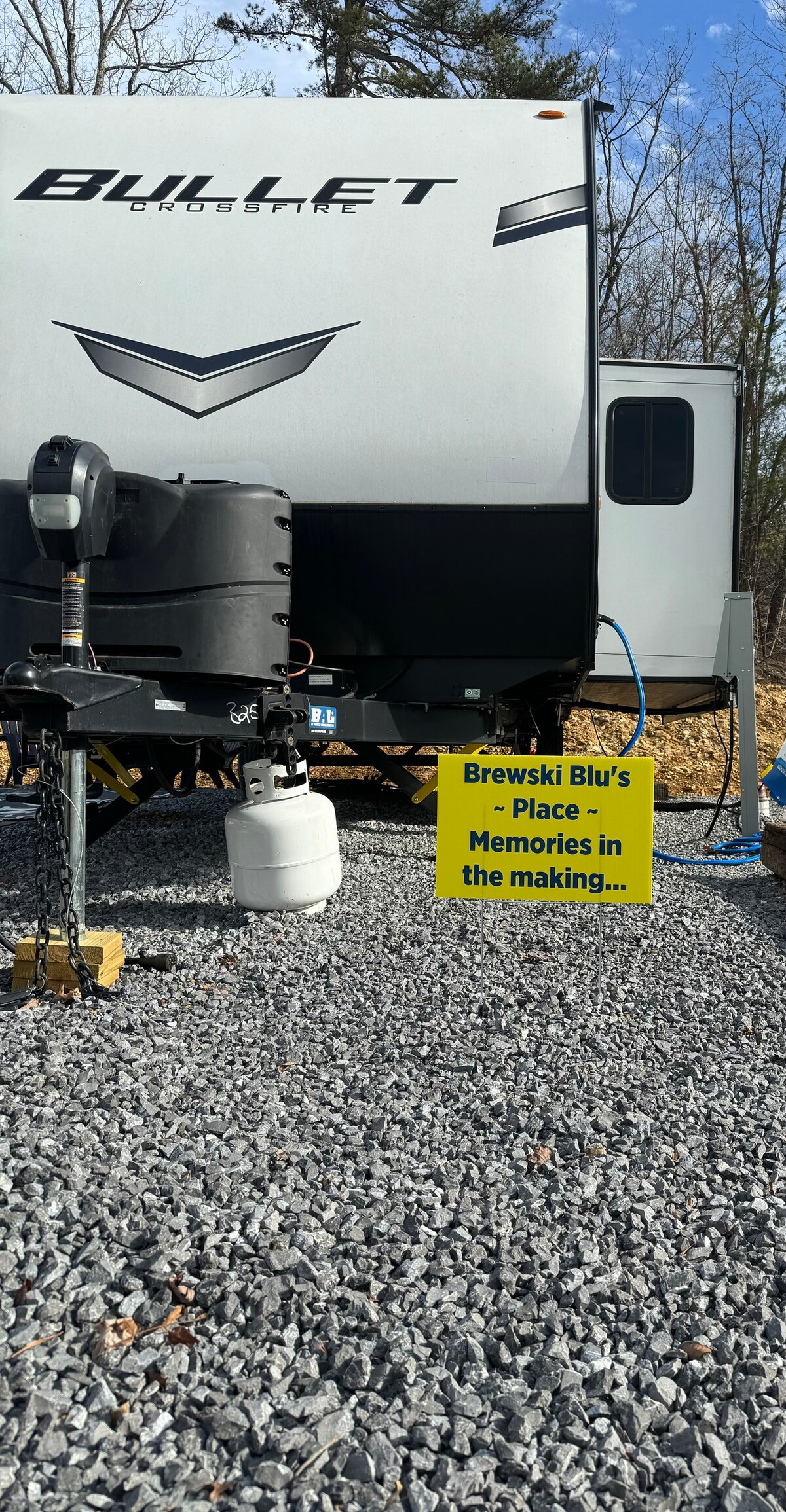 A newly designed camper stands amidst a gravel area, showcasing a smooth exterior with a streamlined shape. Nearby, a propane tank and water hookup are visible, indicating readiness for camping activities. The surrounding landscape features trees and an open sky.