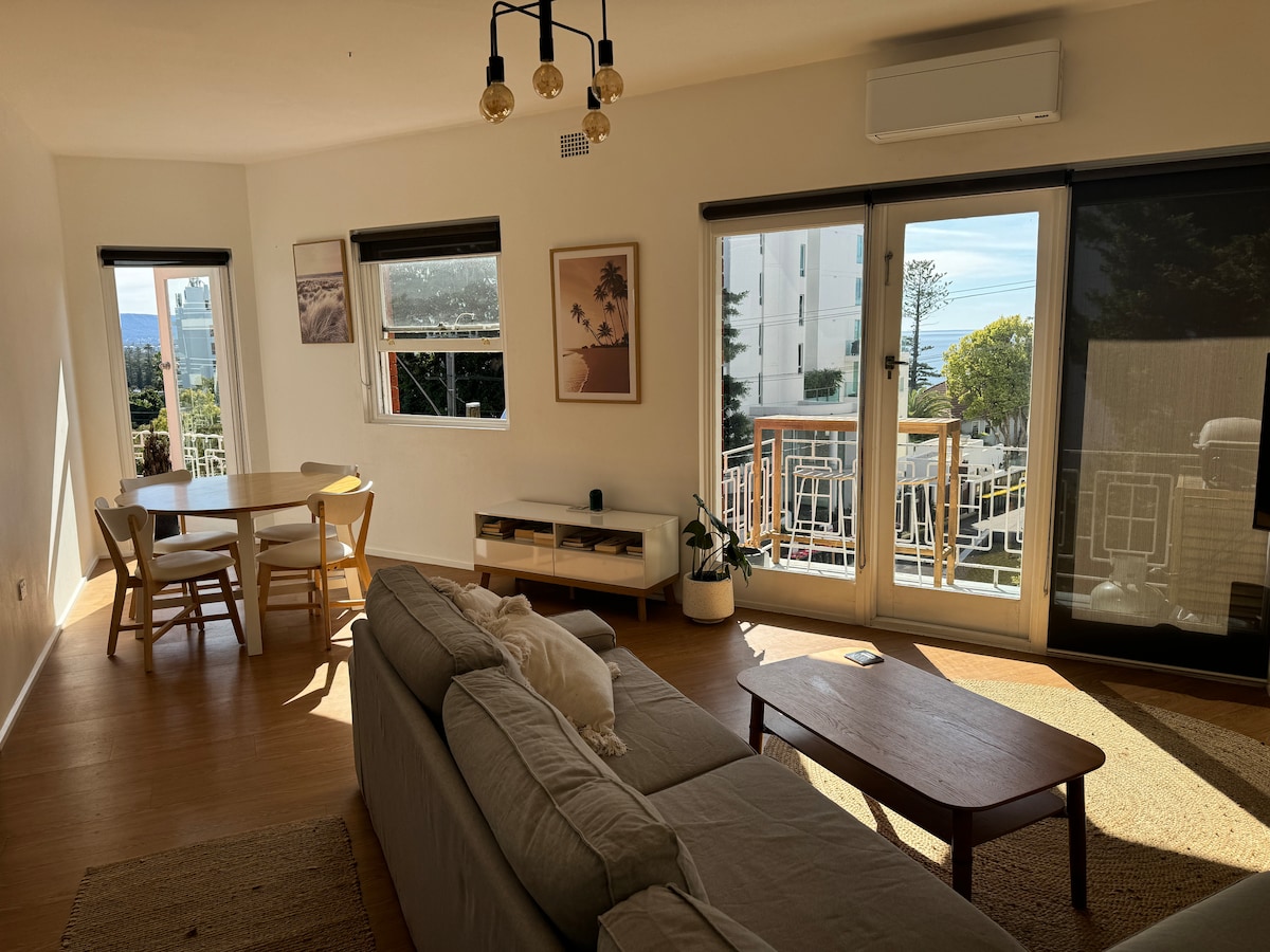 A bright living area is showcased with large windows allowing natural light. A comfortable sofa is positioned in the center, complemented by a wooden coffee table and a dining set for four. Sliding glass doors lead to a balcony, enhancing the open-air feel.