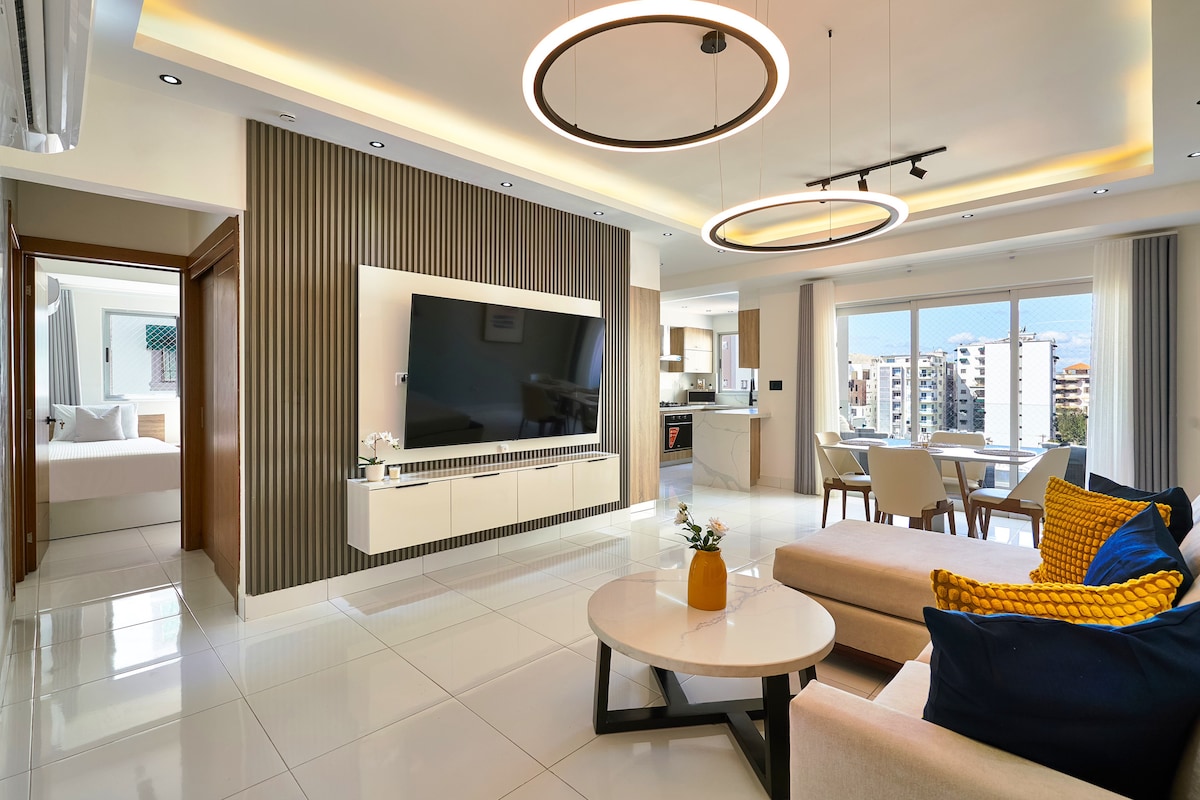 A bright living area features modern furnishings and a large flat-screen television mounted on a stylish wall. Natural light fills the space through expansive windows, which offer views of the city. A cozy seating arrangement includes neutral-colored sofas, accented by colorful throw pillows.