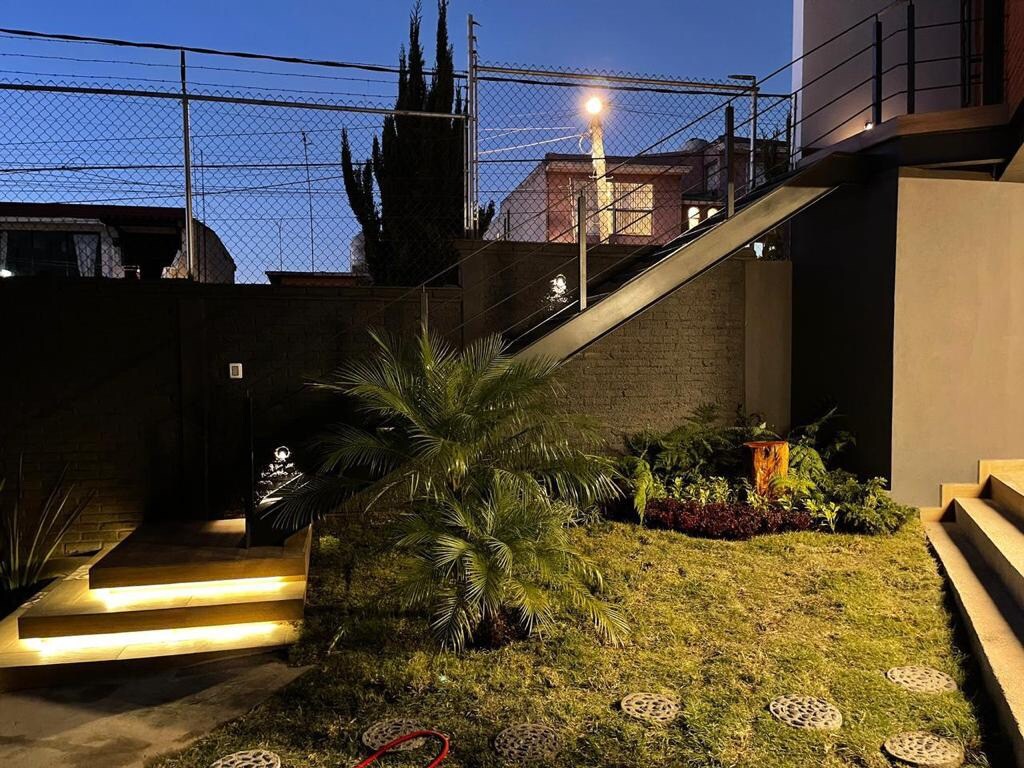 An outdoor area is illuminated by soft lighting, showcasing a manicured garden with greenery and decorative plants. A staircase is visible, leading to an elevated entrance. The surroundings present a modern aesthetic, creating a welcoming atmosphere as evening approaches.