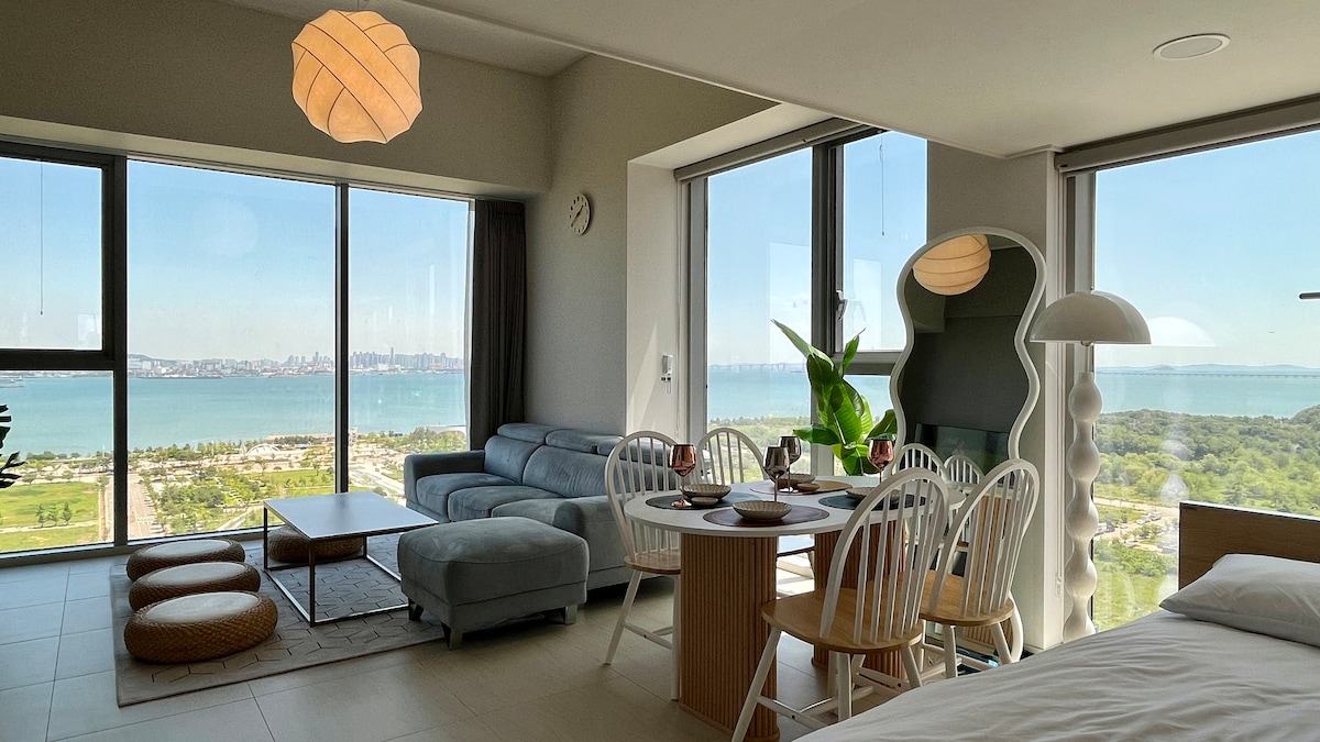 A living area features large floor-to-ceiling windows offering views of the ocean and nearby park. A light-colored sofa and a dining table with white chairs are positioned in the space. A modern lamp and potted plant add a touch of greenery.