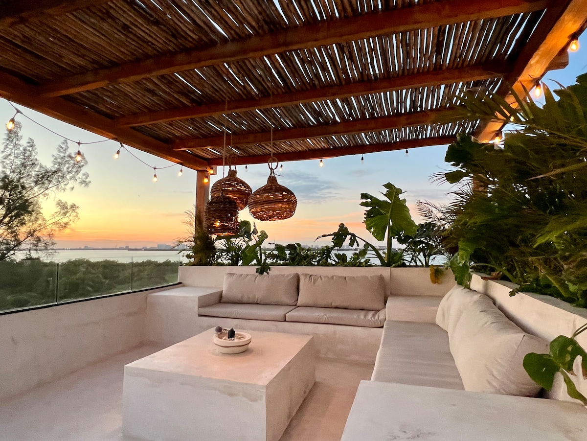 A private rooftop lounge area is surrounded by lush greenery, featuring a spacious sectional sofa and a low coffee table. The space is illuminated by pendant lights, with a scenic view of the sunset reflecting off the water.