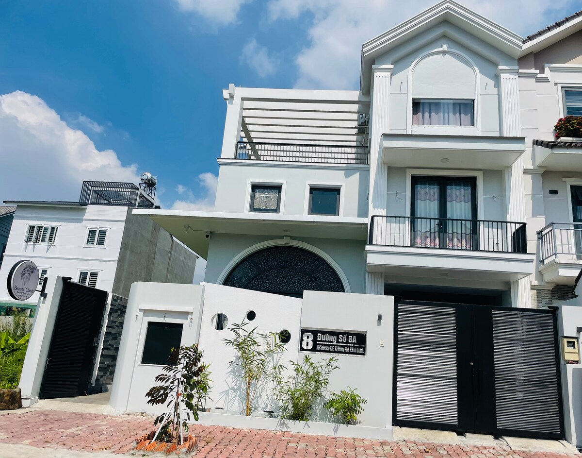 A modern two-story building is featured, with a striking façade incorporating large arched windows. The structure is painted in soft tones, complemented by balconies on the upper level. A neatly paved pathway leads to the entrance, surrounded by minimal landscaping.
