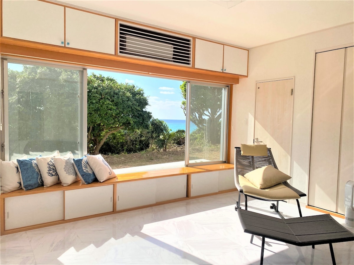 A sunlit interior space features large windows that frame views of lush greenery and the distant ocean. A minimalist seating area includes a cozy sofa adorned with blue and white cushions, complemented by a sleek chair positioned for relaxation.