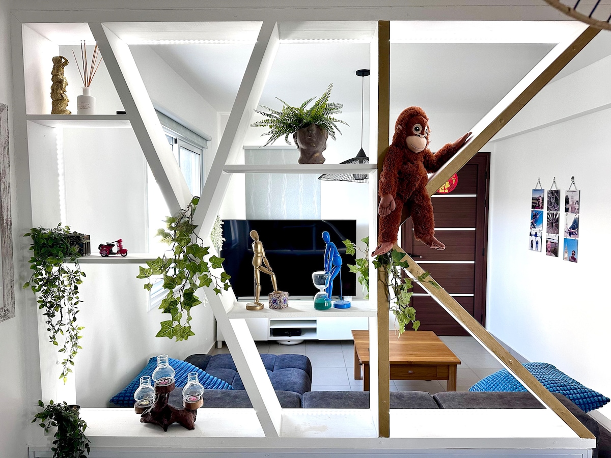 A modern living area is featured, characterized by a stylish open layout with a wooden frame dividing the space. A cozy sofa set provides seating, and decorative shelves display plants and various objects. A television is positioned prominently for entertainment.