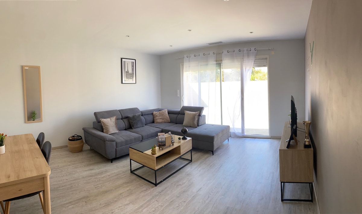 A spacious living area is featured with light wood flooring and neutral-colored walls. A gray sectional sofa occupies one corner, complemented by a low coffee table. A dining table is positioned nearby, while large windows allow natural light to fill the room.