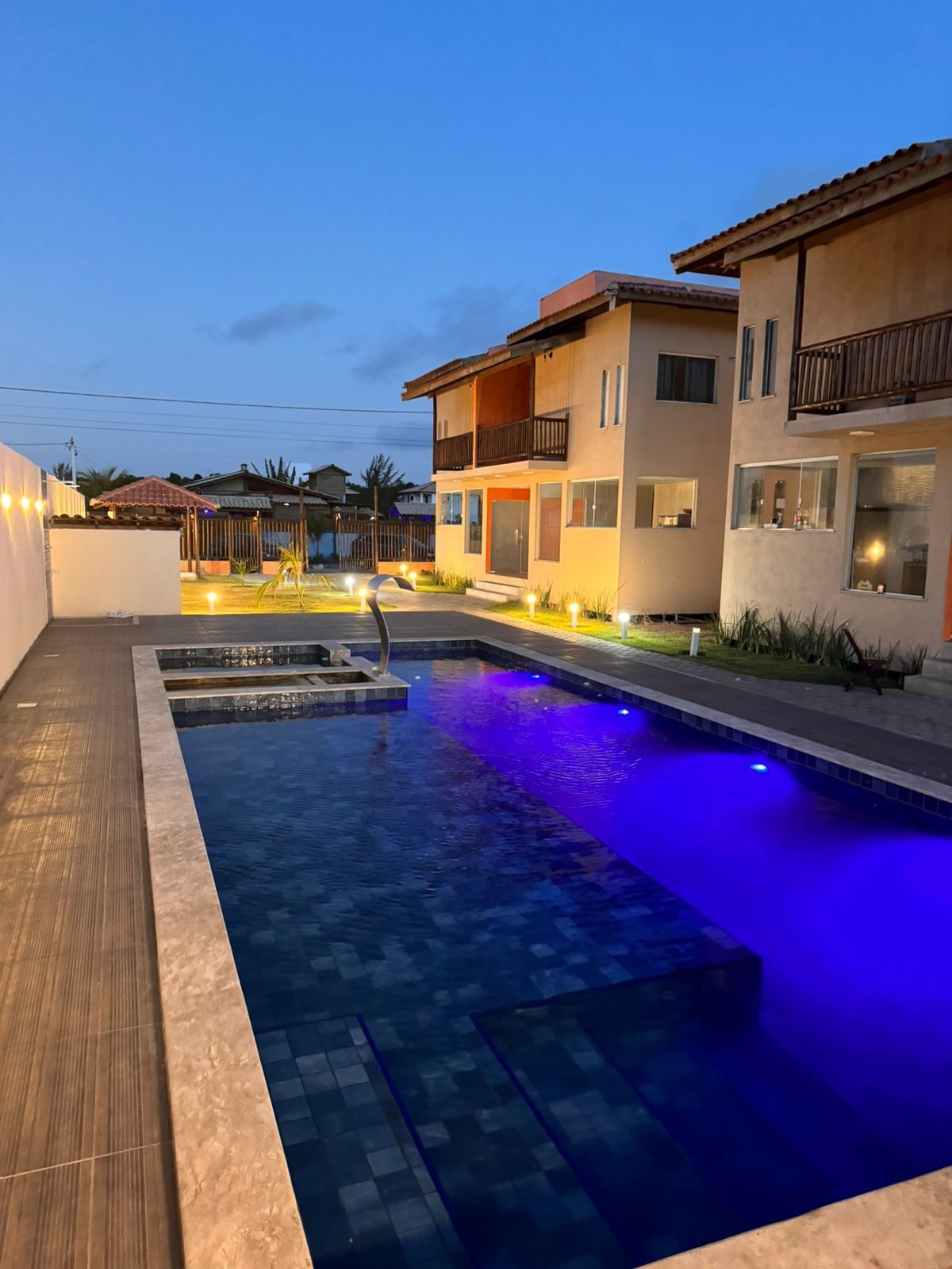 A spacious outdoor area features a large pool illuminated in soft blue light, surrounded by modern buildings. A smaller hot tub is integrated into the pool design. The space is complimented by well-maintained landscaping and warm lighting, creating a serene evening ambiance.