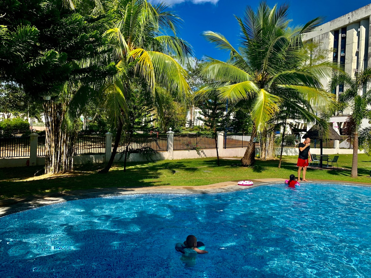 A serene outdoor area features a sparkling pool surrounded by lush palm trees. Children can be seen enjoying the water while an adult stands nearby. The clear blue sky complements the tranquil setting, creating an inviting space for relaxation.