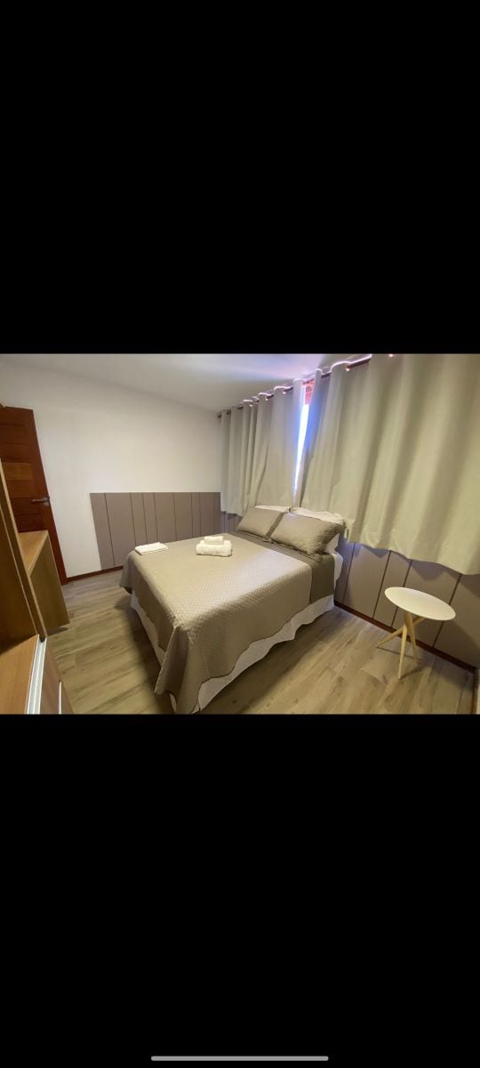 A comfortable sleeping area is presented with a spacious bed, covered in neutral-toned bedding. Light filtered through the partially closed curtains brightens the room. A small side table complements the arrangement, while built-in cabinetry offers additional storage.
