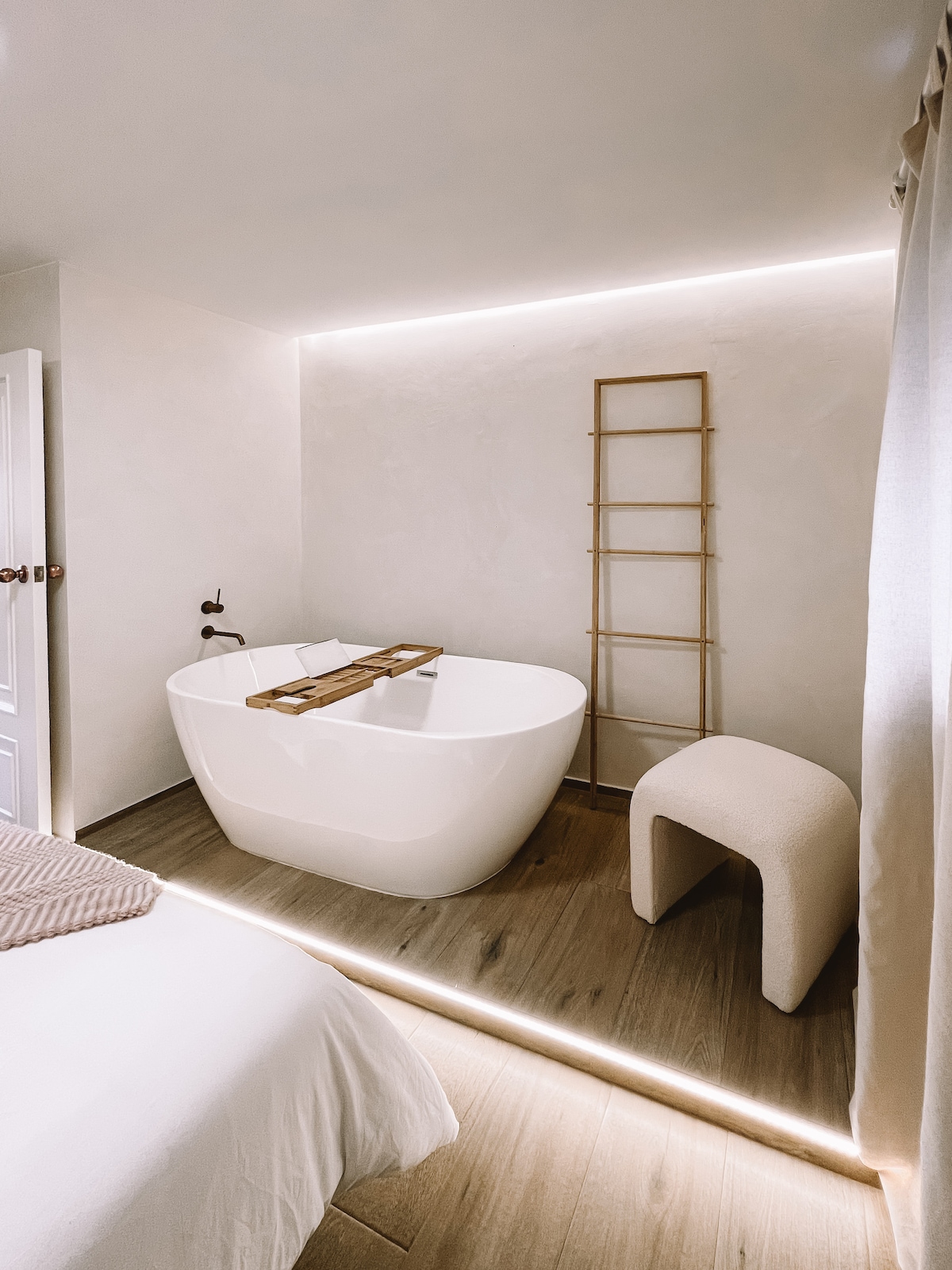 A modern bathroom features a freestanding bathtub, accented by soft, ambient lighting. A wooden bath tray rests on the edge of the tub. A sculptural stool is positioned nearby, with a light-colored curtain providing subtle texture in the space.