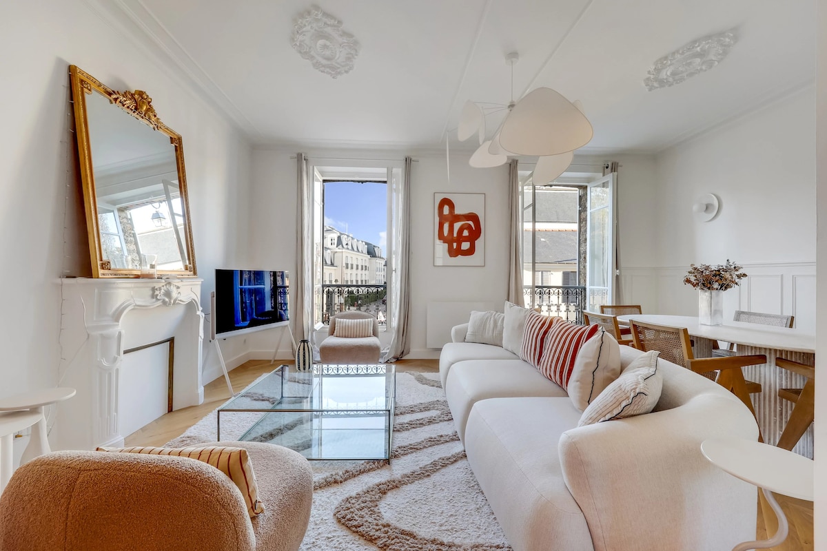 A bright living area features a spacious white sofa with patterned cushions and a glass coffee table. Two sets of French doors lead to a balcony, allowing natural light to fill the space. A dining table sits nearby, accompanied by contemporary lighting fixtures.