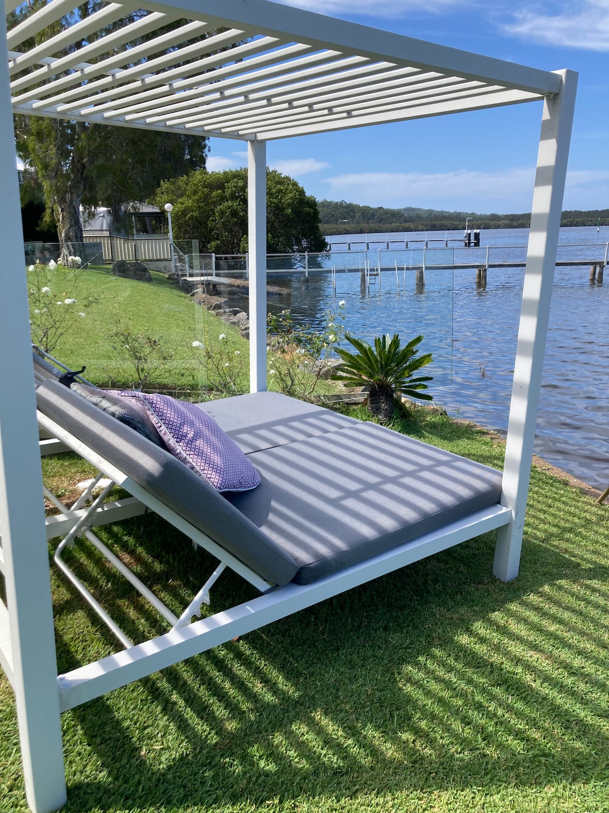 A shaded outdoor lounge area features a comfortable grey cushion under a slatted canopy. The surrounding grass is well-kept, with a small plant nearby. The water reflects the blue sky while a wooden jetty extends into the distance, providing a scenic view across the waterfront.