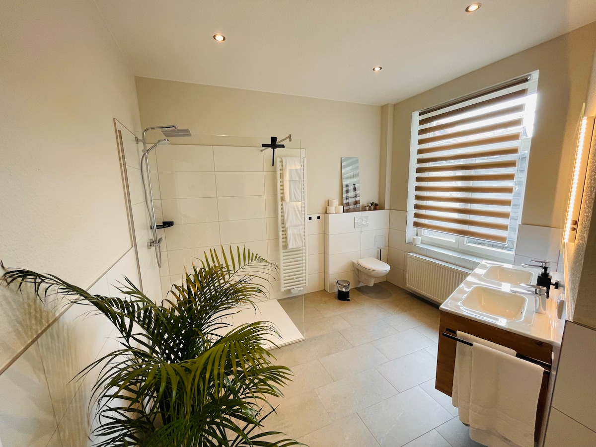 A spacious bathroom is equipped with a walk-in shower, sleek vanities, and modern fixtures. Natural light filters through large windows adorned with striped blinds. A green plant adds a touch of nature, complementing the clean and contemporary design elements throughout the space.