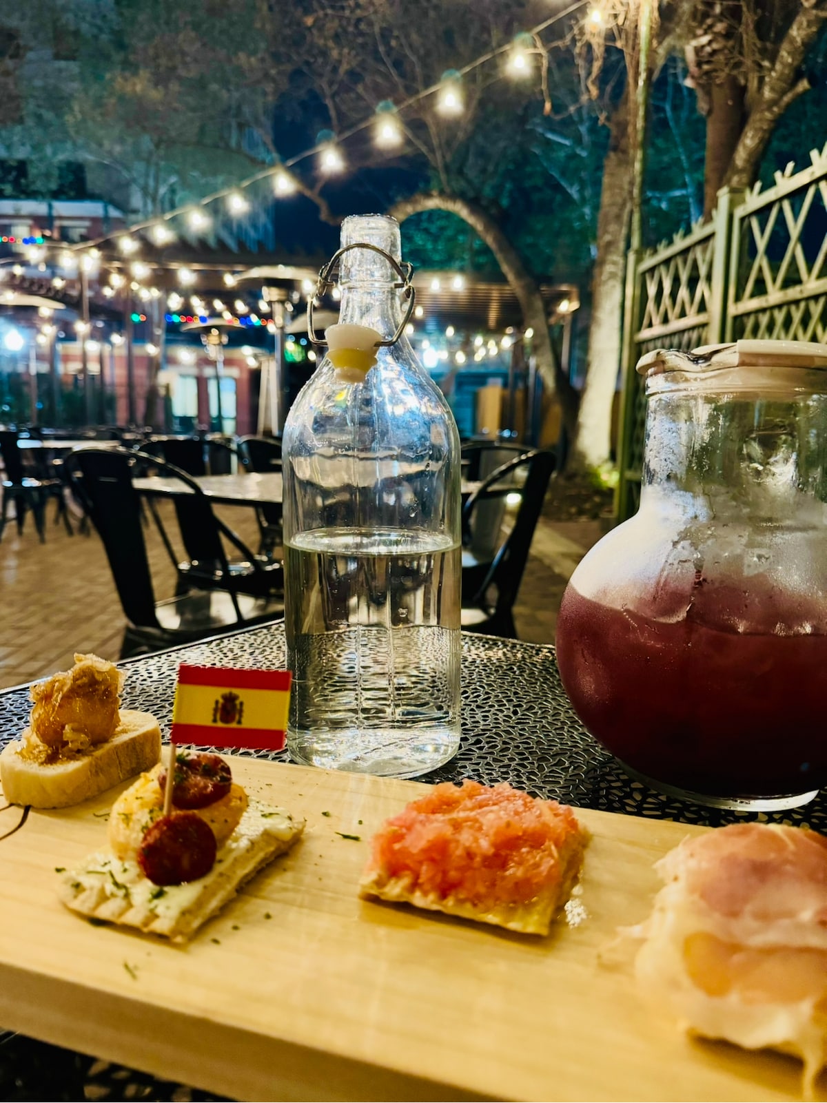 A wooden serving board presents a variety of Spanish tapas, accompanied by a glass pitcher of sangria and a bottle of water. Soft lights twinkle in the background amidst trees, enhancing the outdoor dining setting. A small flag representing Spain is also displayed.