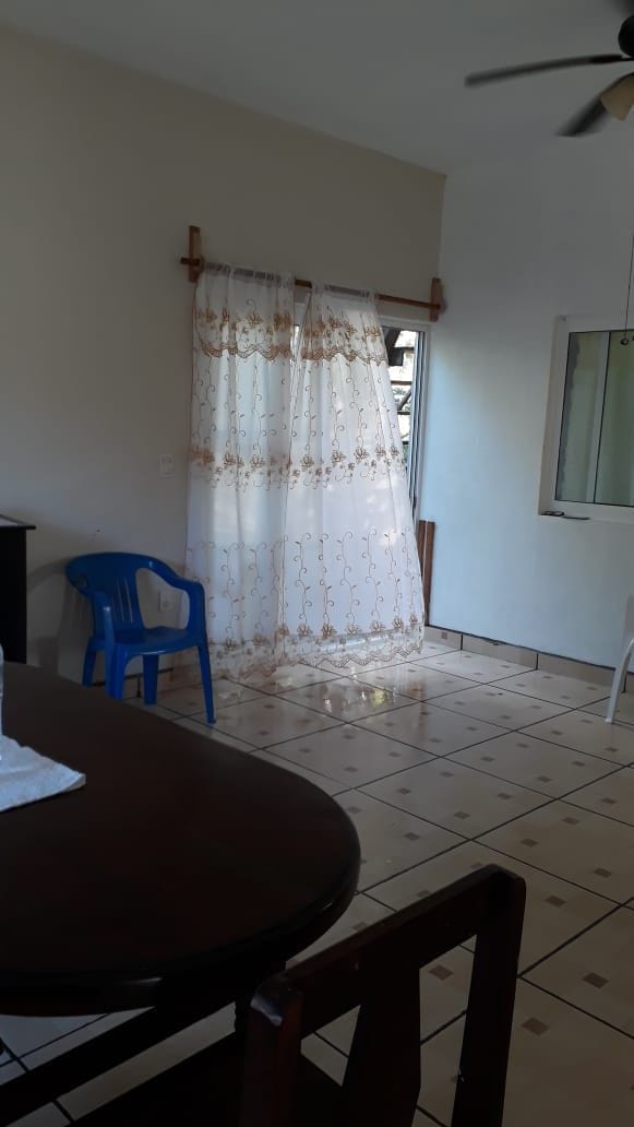 A bright and airy living space features a blue plastic chair beside a wooden dining table. Elegant curtains, adorned with a simple pattern, hang beside a window allowing natural light to filter in. Ceramic tile flooring enhances the open atmosphere of the room.