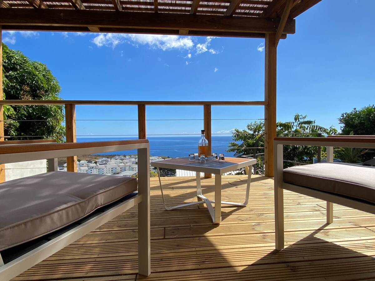 A wooden deck is presented with two comfortable chairs facing a small table. The ocean is visible in the background, under a clear blue sky. Greenery surrounds the space, providing a relaxing outdoor atmosphere.