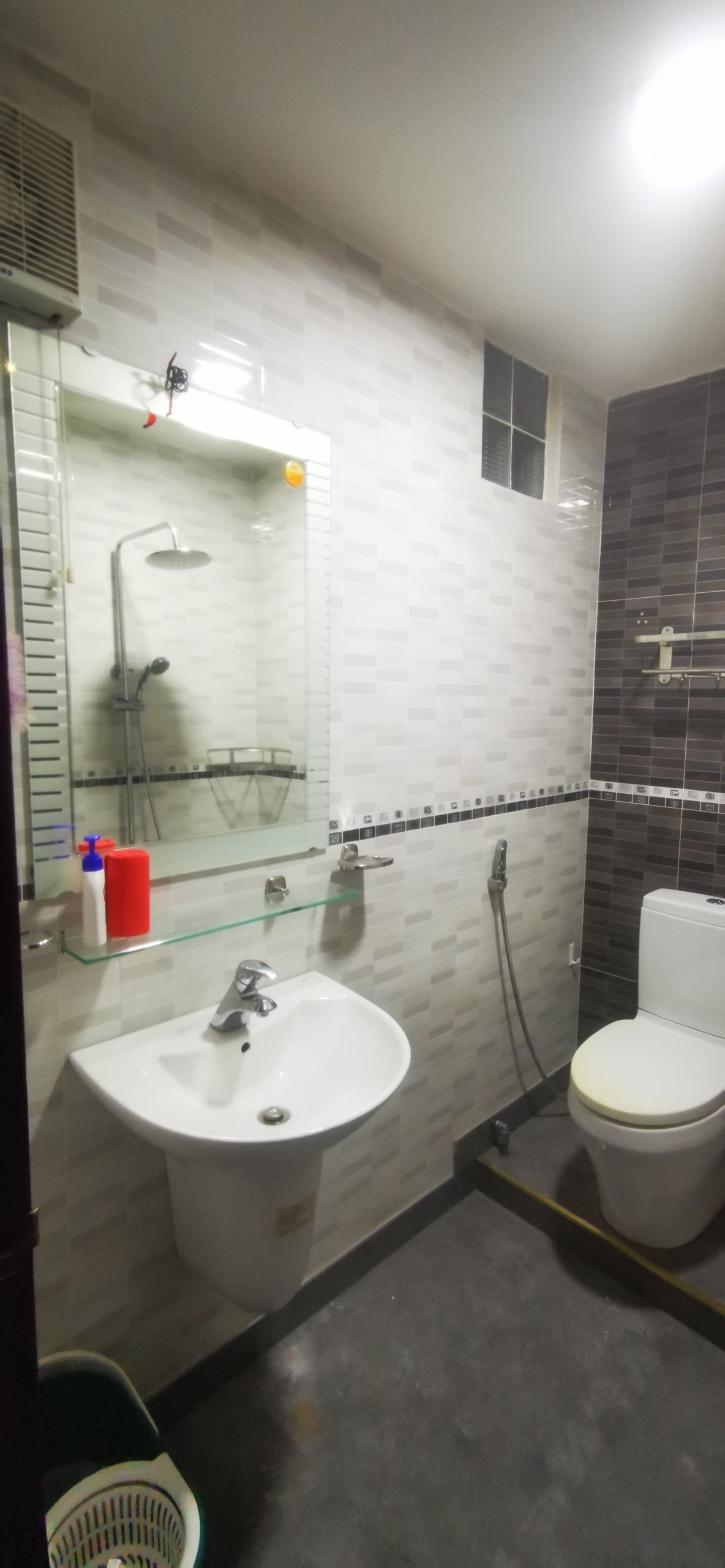 A modern bathroom is presented with a wall-mounted sink and a toilet. A mirrored shelf above the sink reflects the space. The shower area features a glass partition, and the walls are tiled in a combination of light and dark patterns. Ample brightness is provided by overhead lighting.
