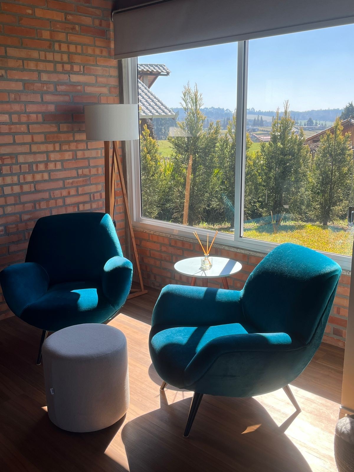 Two plush teal chairs are positioned beside a small round table, creating a cozy seating area. A modern lamp stands nearby, and natural light floods in through large windows, offering views of the surrounding greenery and distant hills.