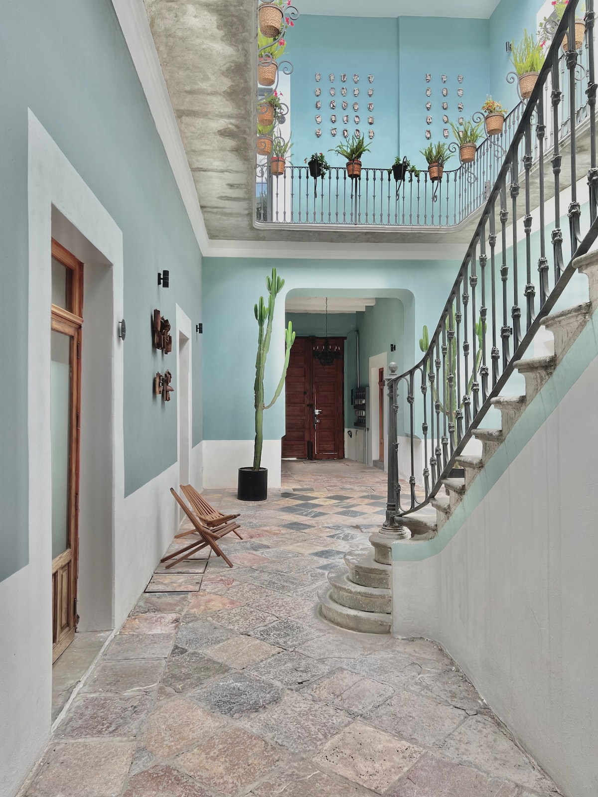 A hall features a decorative staircase with a wrought iron railing and soft blue walls. Potted plants are arranged along the balcony above, and a wooden chair is positioned on the textured stone floor, creating a tranquil entryway with a blend of original and modern elements.
