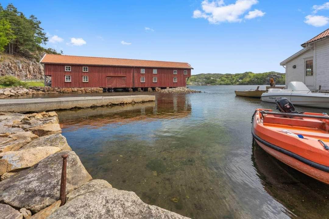 The image showcases a calm waterfront scene featuring a red boathouse with multiple windows adjacent to the water. Two boats, one orange and one white, are moored at the shoreline, with rocky elements lining the dock. Lush greenery is visible in the background.