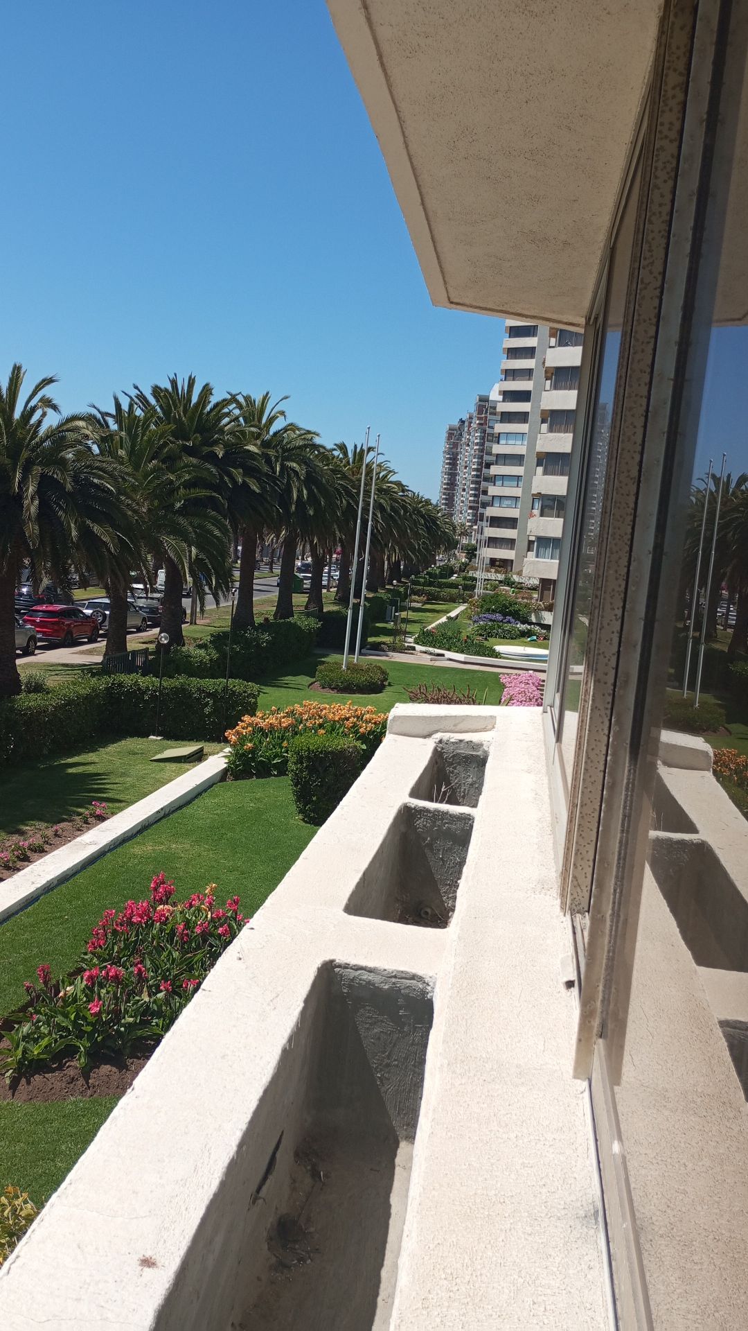 A balcony view reveals a landscaped area with palm trees and floral arrangements. The well-maintained garden features vibrant flowers and a walkway leading toward nearby buildings. Sunlight casts a bright, inviting atmosphere across the scene.