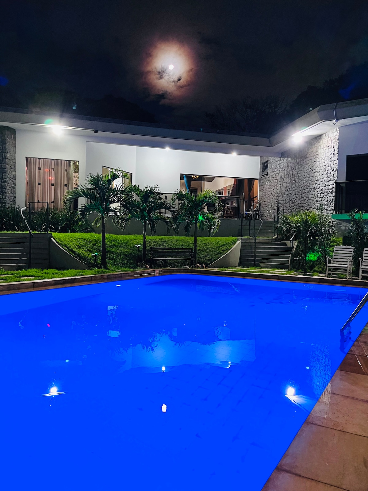 A well-lit swimming pool with sparkling blue water is framed by lush greenery and landscaped steps. The exterior of the house is visible in the background, showcasing large windows that reflect the warm lighting of the space. The atmosphere appears tranquil and inviting at night.