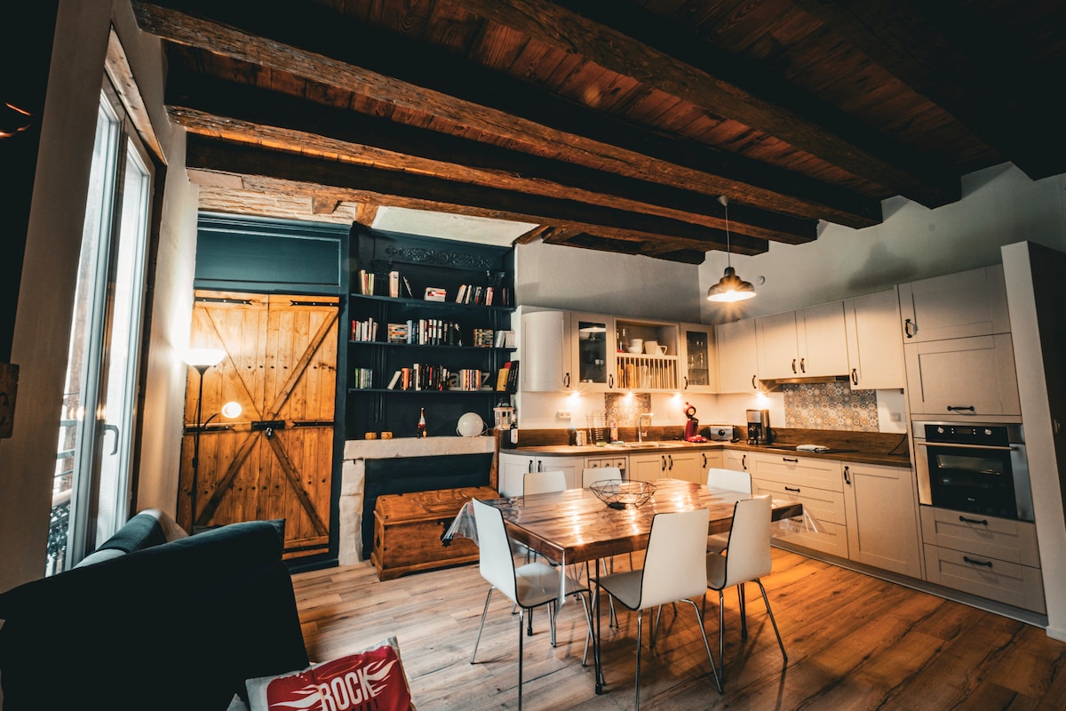 An open-concept kitchen and dining area features wooden beams overhead, a rustic wooden door in the background, and a modern kitchen with white cabinetry. A dining table is surrounded by white chairs, with a soft seating area visible in the corner.