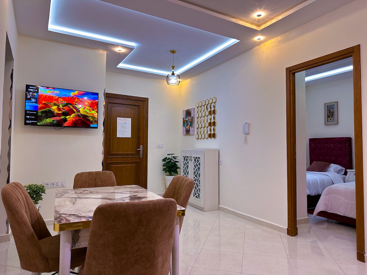 A bright and modern living space features a marble dining table surrounded by four upholstered chairs. A flat-screen television is mounted on the wall, and an elegantly framed door leads to a well-appointed bedroom. Soft lighting adds a welcoming touch to the area.