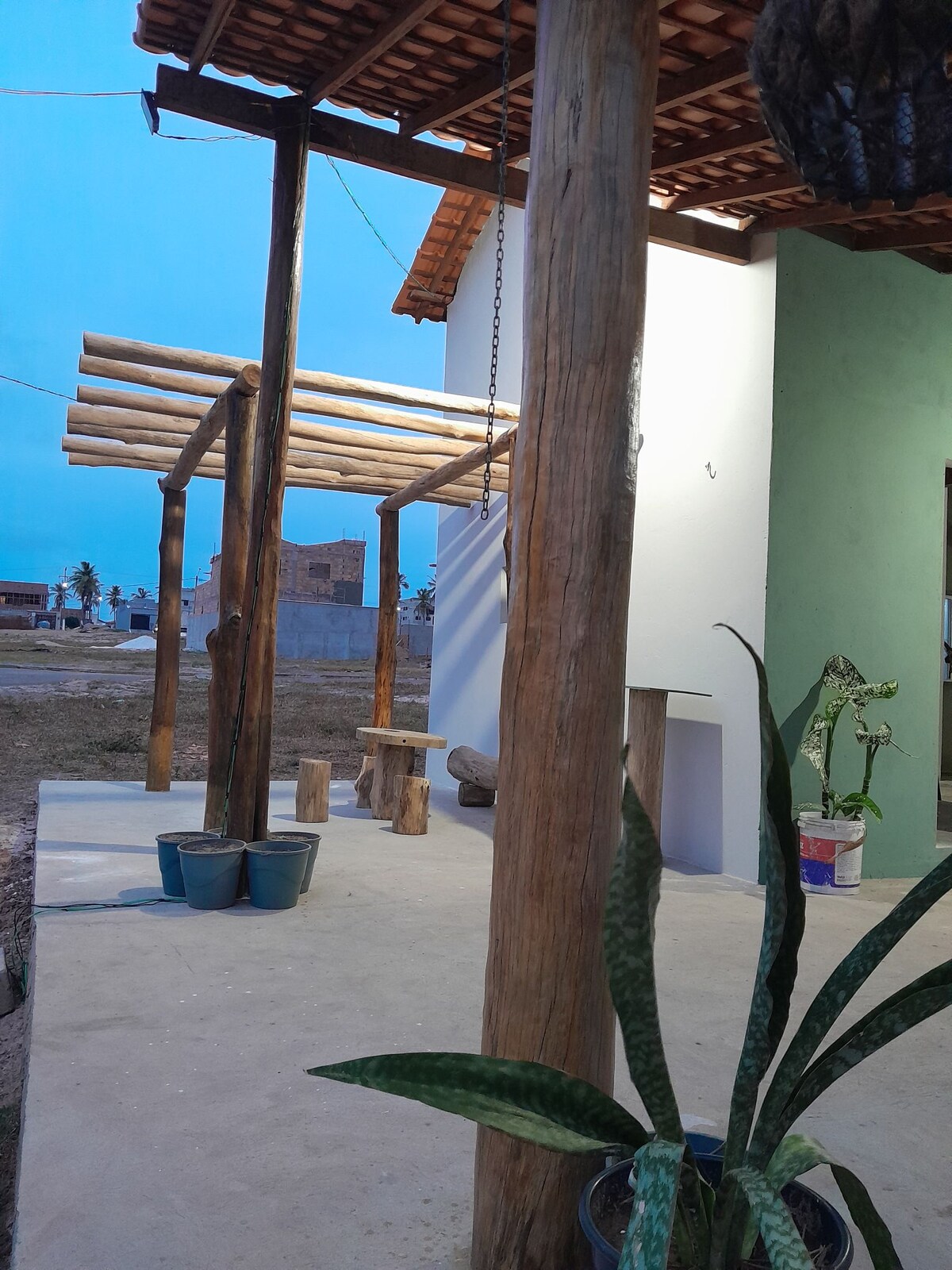 An outdoor area features a rustic wooden structure supported by tree trunks, providing shade. A simple wooden bench is positioned nearby, with potted plants arranged on the ground, adding a touch of greenery. The scene is set against an early evening sky.