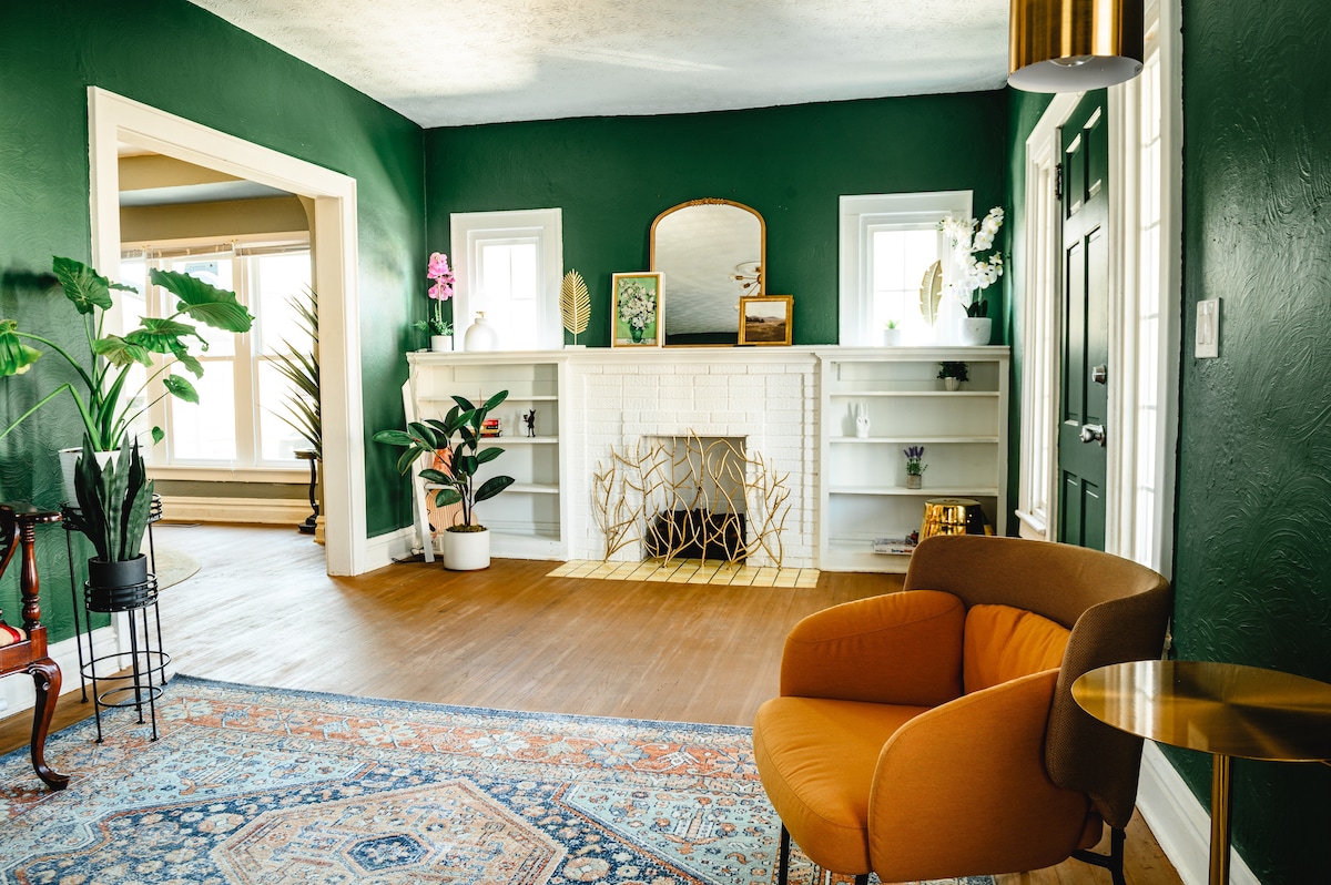 A spacious living area features deep green walls and hardwood flooring. A decorative fireplace with a gold accent frame is set against a white mantel. A cozy orange armchair is positioned next to a small side table, with plants adding a touch of greenery to the space.