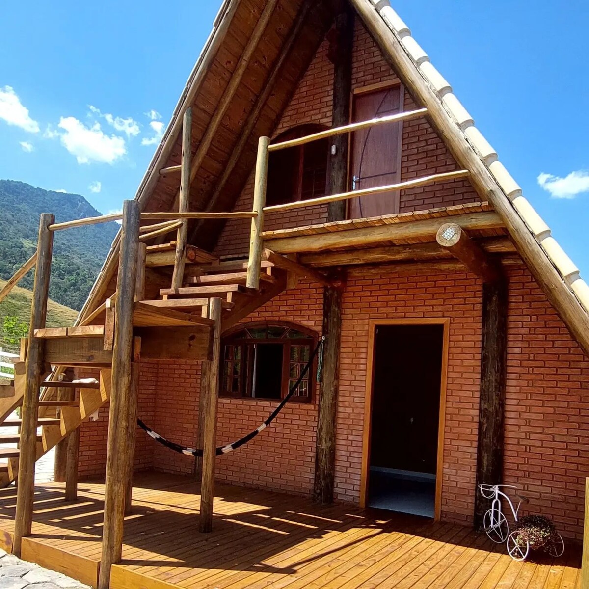 A wooden cabin features a unique triangular design with a sloped roof and spacious wooden deck. Stairs lead to a second level, and a hammock is suspended on the porch. Surrounding mountains provide a scenic backdrop, enhancing the cabin's rustic charm.