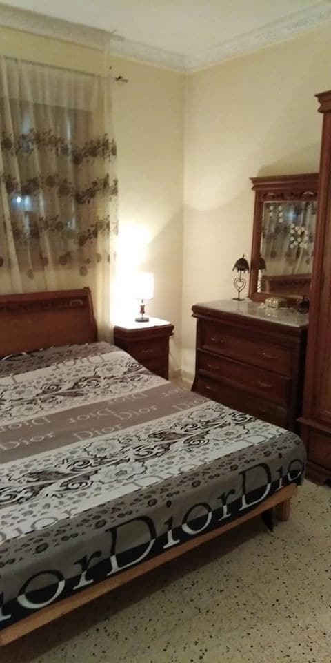 A calming bedroom features a spacious bed adorned with a patterned quilt. A bedside lamp provides soft lighting next to the bed. Antique furniture, including a dresser and a wardrobe, complements the decor. Light drapes softly over the window, enhancing the warm tones of the room.