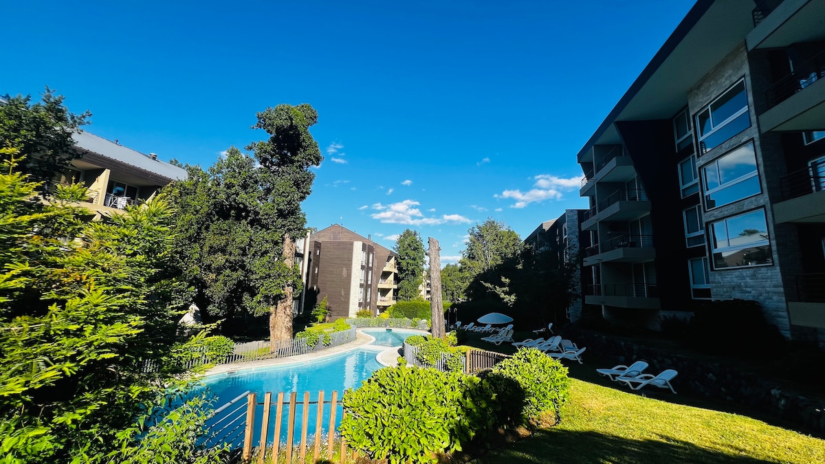 A serene outdoor area features a winding pool surrounded by lush greenery and well-maintained landscaping. Several lounge chairs are positioned near the water, providing options for relaxation. Adjacent buildings are visible, offering a glimpse of the overall property layout.