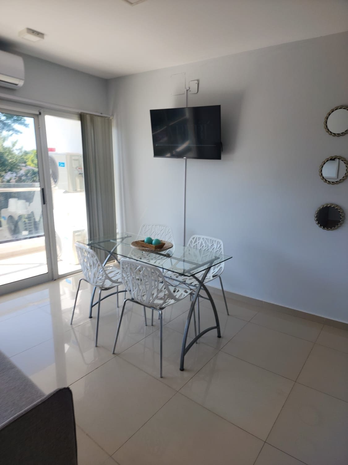 A modern dining area is visible, featuring a glass table surrounded by four transparent chairs. A wall-mounted television is positioned above the table, and two circular mirrors are displayed on the wall. Natural light enters through sliding glass doors, leading to an outdoor space.