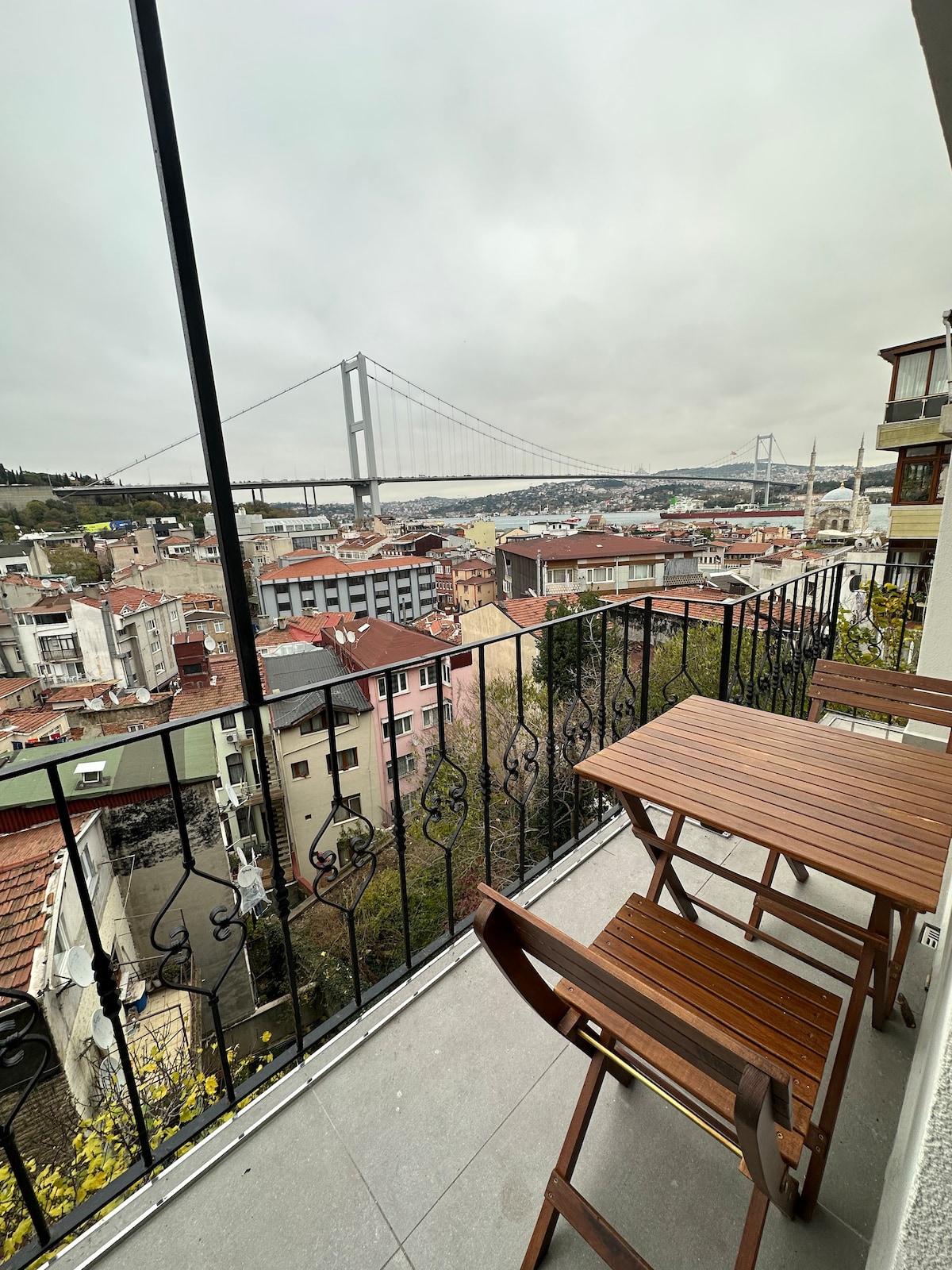 A private balcony is equipped with a wooden table and chairs, offering a view of the city below and a distant bridge. The surrounding area showcases a mix of buildings and greenery, creating a peaceful outdoor space.