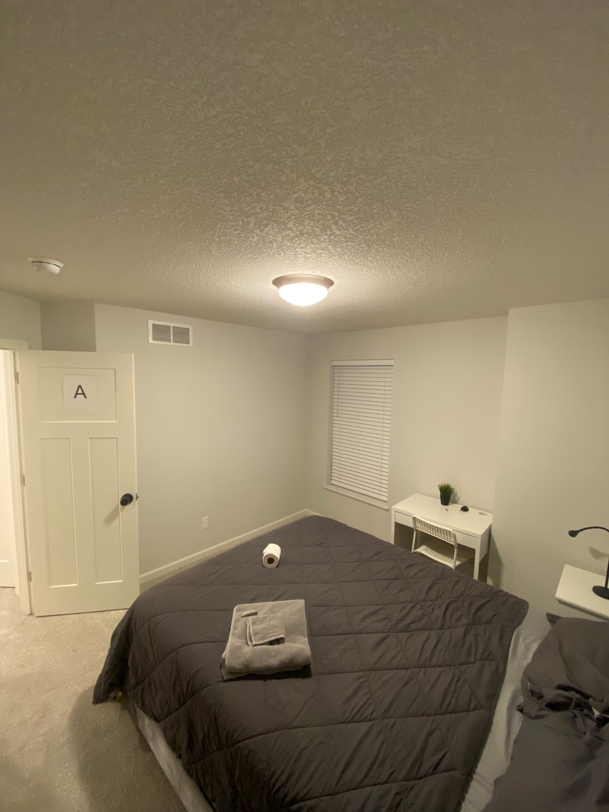 A private room features a spacious bed covered with a dark quilt, complemented by a neatly folded towel. A small bedside table with a lamp stands next to the bed. Natural light enters through a window with white blinds, while a closet door is visible in the background.
