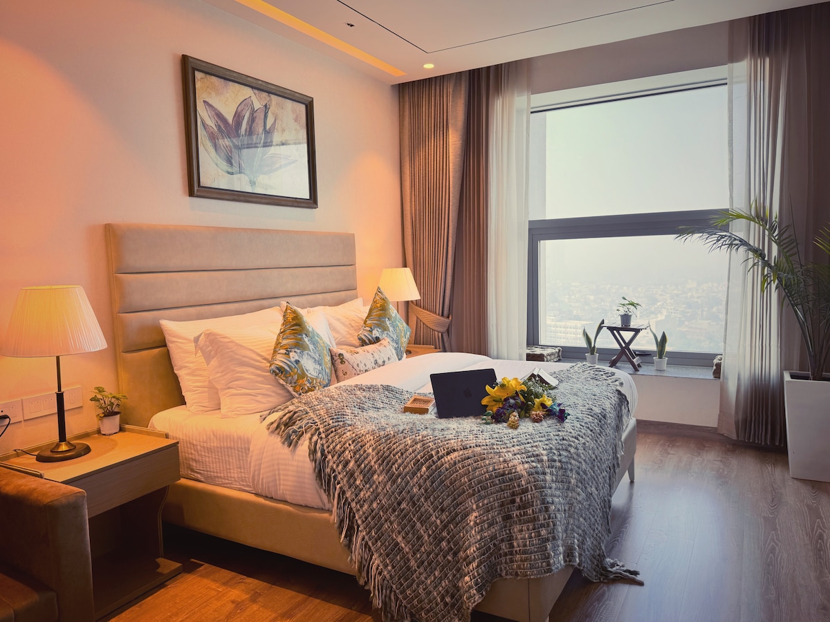 A spacious bedroom features a large bed adorned with decorative pillows and a textured throw blanket. Two bedside lamps provide soft lighting. A large window offers a view of the skyline, complemented by a small table with plants and fresh flowers.