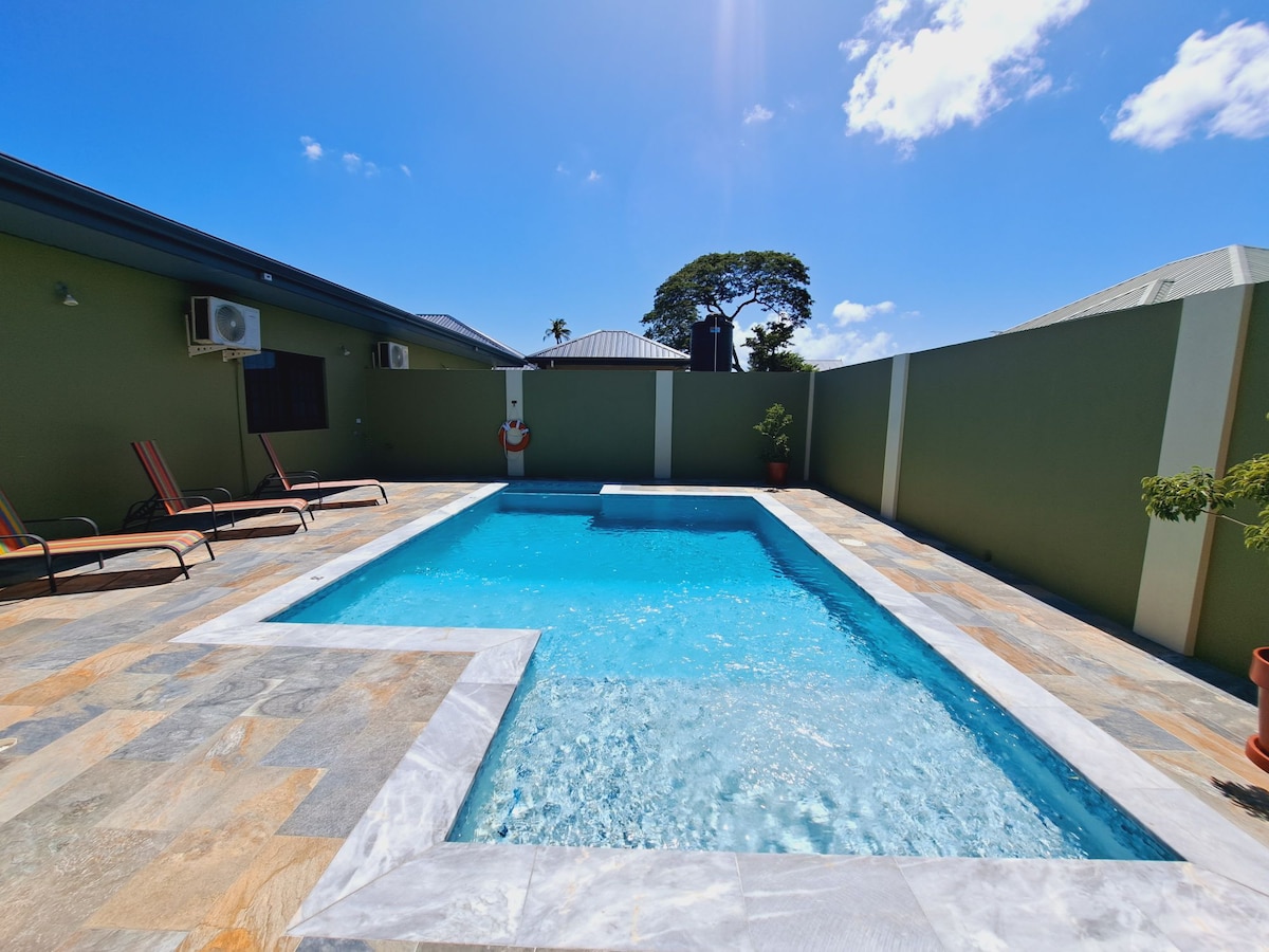 A private swimming pool is featured in a tranquil outdoor area, surrounded by a stone patio. Sun loungers are positioned near the pool, with greenery along the perimeter. The clear blue water of the pool reflects the bright blue sky above.