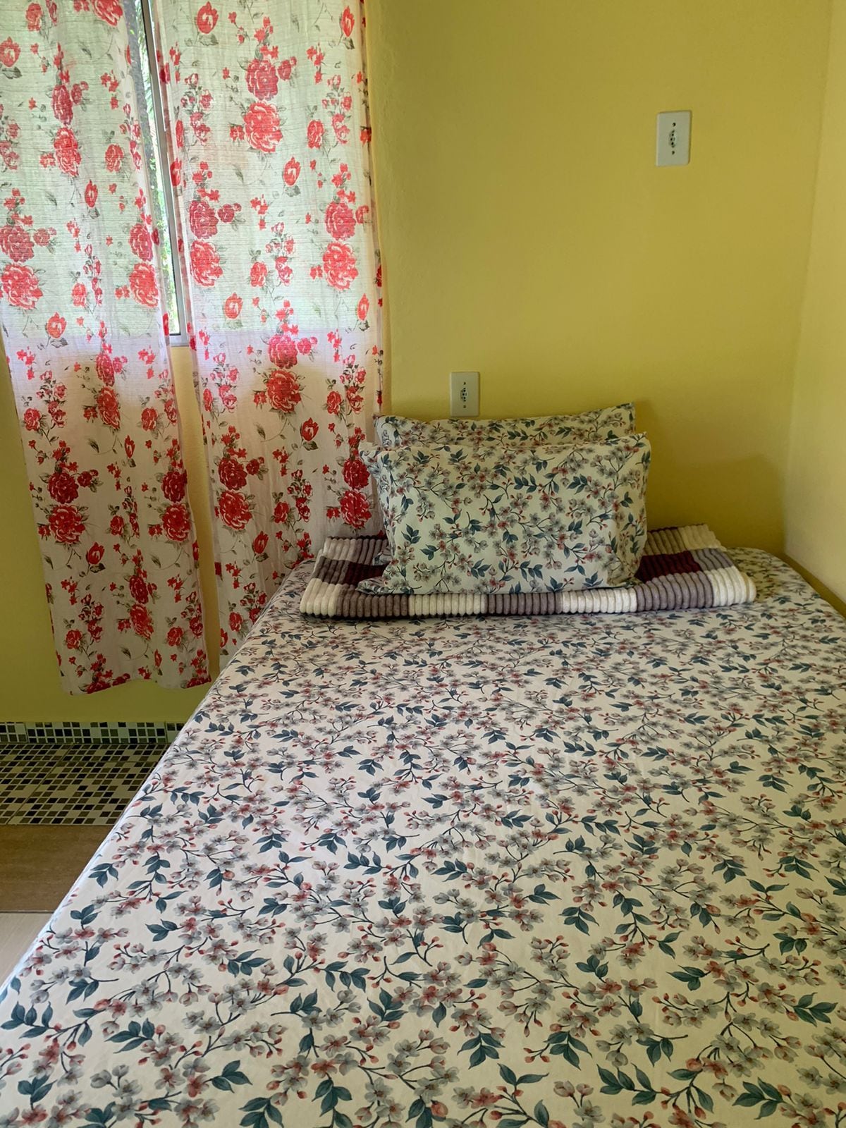 A single bed is dressed in a floral-patterned coverlet, complemented by a matching pillow and decorative cushion. Yellow walls create a warm ambiance, while sheer floral curtains gently frame the window, allowing natural light to brighten the space.