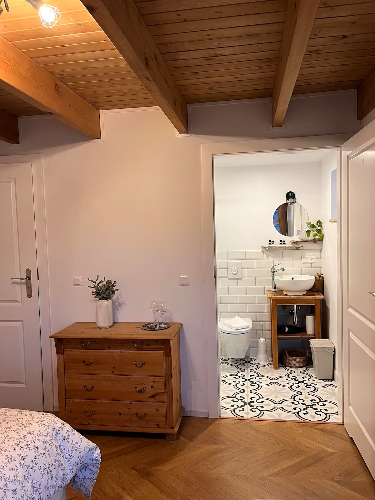 A light and airy room features wooden beams and warm-toned flooring. A dresser made of natural wood sits near the entrance, while a doorway leads to an ensuite bathroom with a distinct tile pattern. Soft lighting creates a welcoming ambiance.