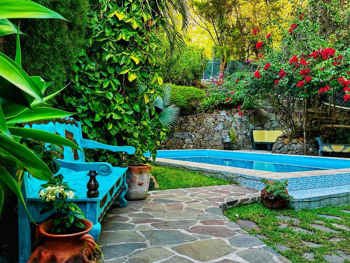 A vibrant blue bench is positioned beside a stone pathway that leads to a heated in-ground pool. Lush greenery and colorful flowering plants surround the area, enhancing the serene atmosphere. The pool area features smooth tiles and natural stone accents.