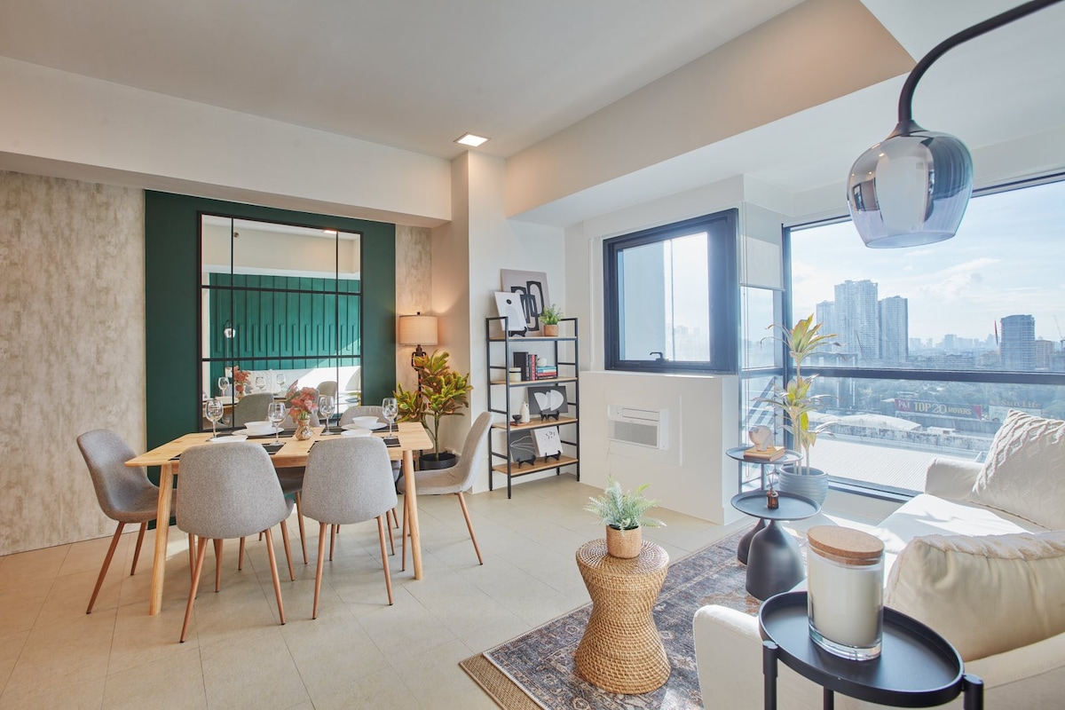 A bright living space features a modern dining area with a wooden table surrounded by four upholstered chairs. A cozy seating area includes a light-colored sofa and a patterned rug. Large windows offer a view of the city skyline, enhancing the natural light within the room.