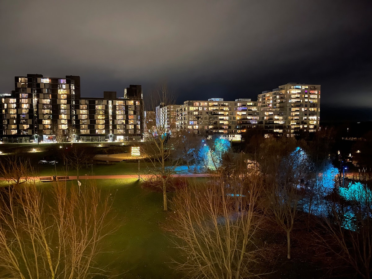 A nighttime view of a modern urban landscape is presented, featuring several multi-story residential buildings illuminated by soft lighting. Trees with bare branches are visible in the foreground, and paths can be seen winding through the landscaped area, enhancing the serene atmosphere.