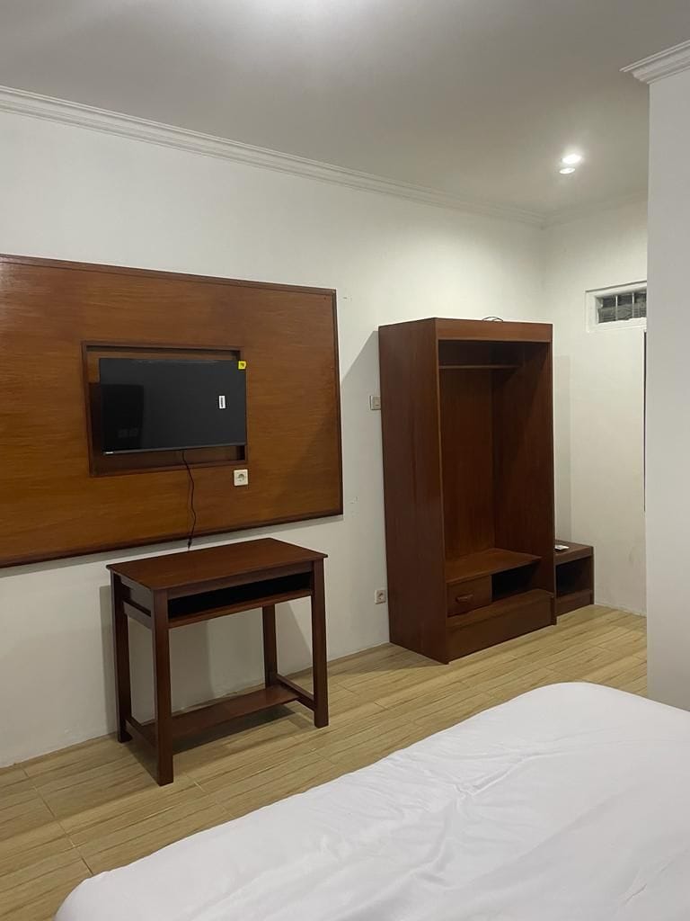 A simple and functional guest room features a wall-mounted television above a wooden desk. A matching wooden wardrobe with shelves and a drawer is positioned nearby. The space is illuminated by ceiling lights and has neutral-colored walls.